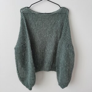 May include: A sage green, oversized sweater with a round neckline and long, voluminous sleeves. The knitwear has a loose, open weave, giving it a soft, airy appearance. The sweater is hanging on a black hanger against a white background.
