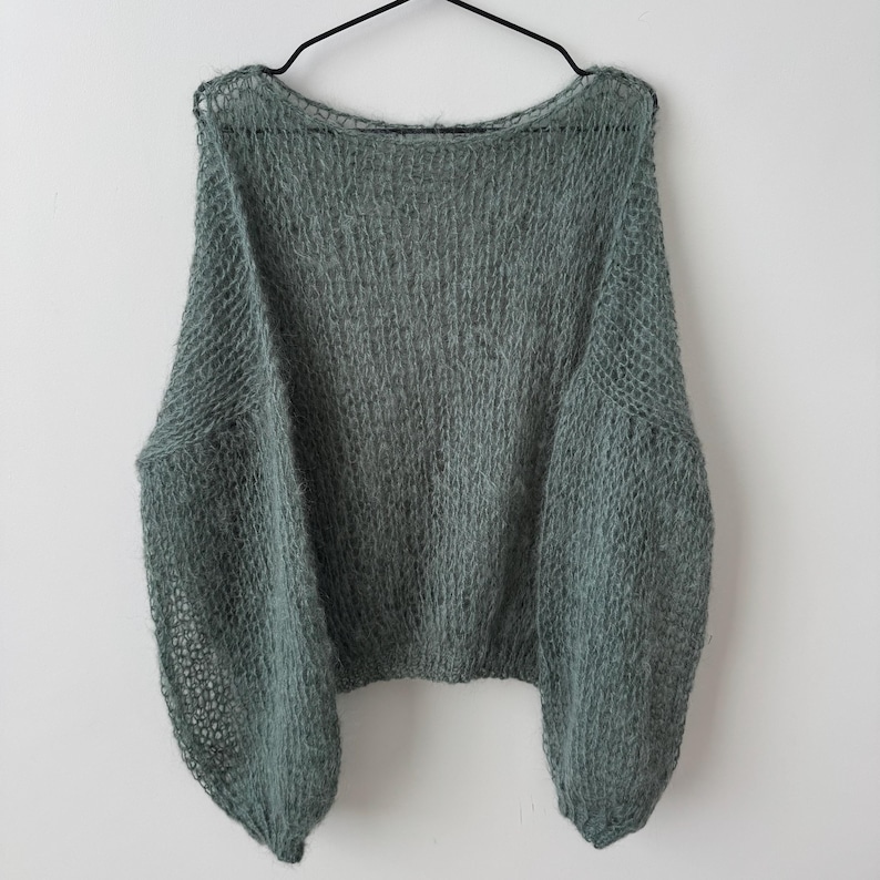 May include: A sage green, oversized sweater with a round neckline and long, voluminous sleeves. The knitwear has a loose, open weave, giving it a soft, airy appearance. The sweater is hanging on a black hanger against a white background.
