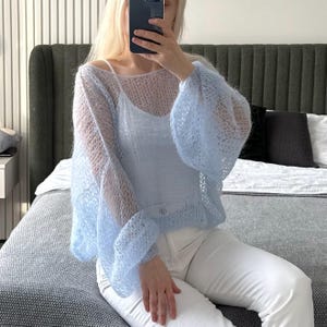Op de afbeelding: Een lichtblauwe, open gebreide trui met wijde mouwen. De trui wordt gedragen over een witte camisole en gecombineerd met een witte broek. De trui heeft een delicate, luchtige uitstraling.