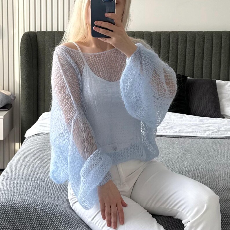 Op de afbeelding: Een lichtblauwe, open gebreide trui met wijde mouwen. De trui wordt gedragen over een witte camisole en gecombineerd met een witte broek. De trui heeft een delicate, luchtige uitstraling.