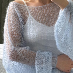 Op de afbeelding: Een lichtblauwe, open gebreide trui met lange mouwen. De trui heeft een delicate, luchtige uitstraling en wordt over een witte camisole gedragen. De mouwen zijn bij de polsen gerimpeld. De textuur van de trui is zacht en licht pluizig.