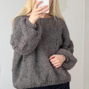 Puede incluir: Un suéter de punto gris de gran tamaño con cuello redondo y mangas largas y voluminosas. El suéter tiene un aspecto ligeramente desgastado con agujeros intencionales. El modelo lleva pantalones negros.