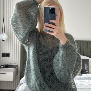 May include: A sage green, open-weave sweater with long, voluminous sleeves. The sweater has a relaxed fit and a delicate, airy texture. The person in the image is holding a phone in front of their face, with blonde hair.