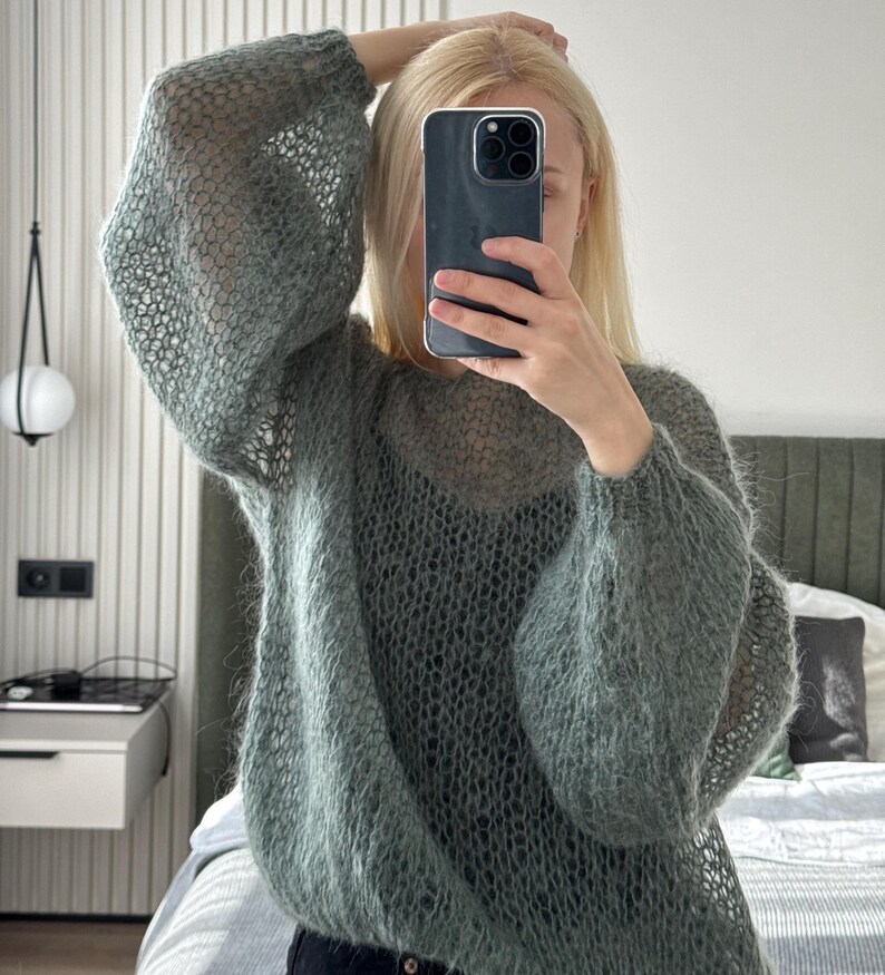 May include: A sage green, open-weave sweater with long, voluminous sleeves. The sweater has a relaxed fit and a delicate, airy texture. The person in the image is holding a phone in front of their face, with blonde hair.