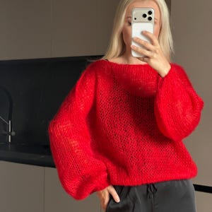 Peut inclure: Un pull rouge vif, ample et à manches longues, confectionné dans un fil duveteux à maille ouverte. Le vêtement présente une large encolure arrondie et des manches volumineuses qui se resserrent aux poignets, offrant une silhouette décontractée.