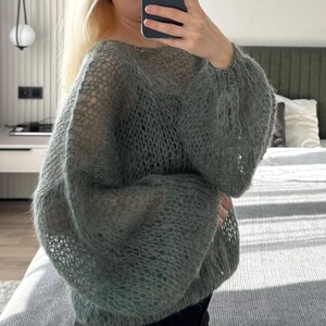 May include: A light green, open-weave sweater with long, voluminous sleeves. The sweater has a loose, airy knit design, and the color is a soft, muted green. The sleeves are gathered at the cuffs, creating a balloon shape. The sweater appears to be made of a soft, possibly fuzzy material.