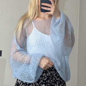 Op de afbeelding: Een lichtblauwe, open gebreide trui met lange, volumineuze mouwen. De trui wordt over een witte camisole gedragen. De onderkant van de afbeelding toont een zwarte rok met bloemenprint. De textuur van de trui is luchtig en delicaat.