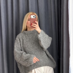 May include: A gray, oversized sweater with a crew neck and long sleeves. The sweater is paired with white pants. The model is holding a phone in front of their face. The background is a gray curtain.