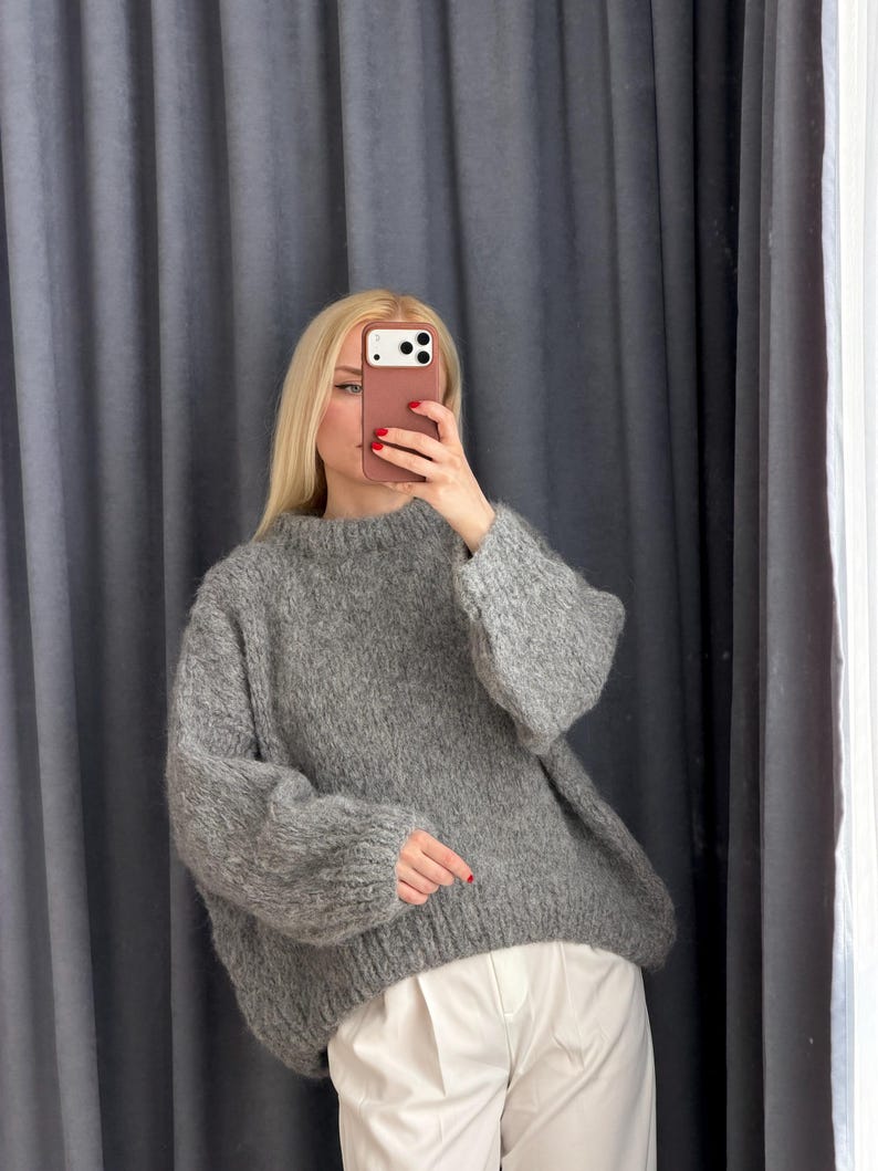 May include: A gray, oversized sweater with a crew neck and long sleeves. The sweater is paired with white pants. The model is holding a phone in front of their face. The background is a gray curtain.