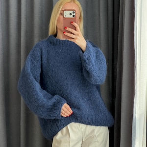 May include: A navy blue, oversized knit sweater with long sleeves. The sweater is paired with light-colored pants. The person in the image is holding a phone up to their face.