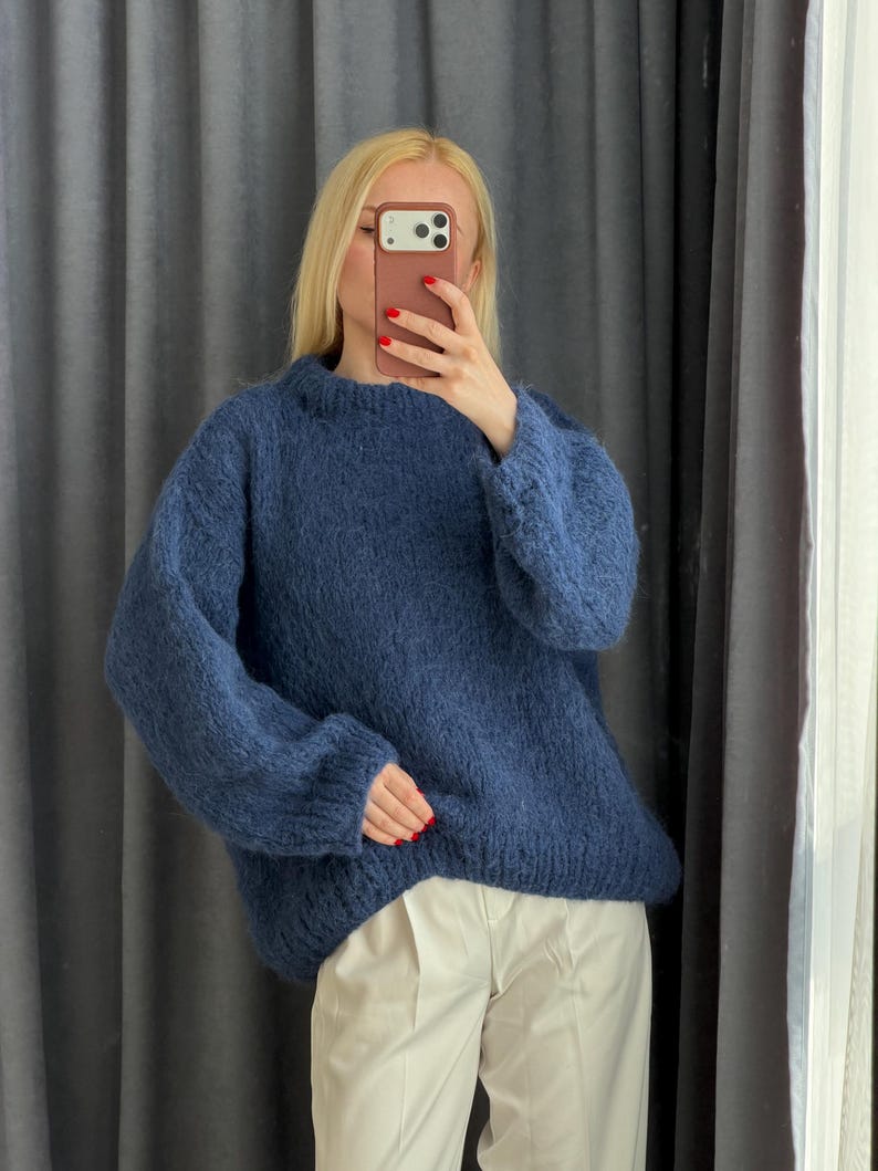 May include: A navy blue, oversized knit sweater with long sleeves. The sweater is paired with light-colored pants. The person in the image is holding a phone up to their face.