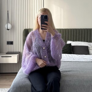 Op de afbeelding: Een doorschijnend, lichtpaars vest met een V-hals en drie parelmoer knopen. De trui met lange mouwen heeft een losse, open weefsel en wordt gedragen met een zwarte broek. Het vest heeft een pastelkleur.