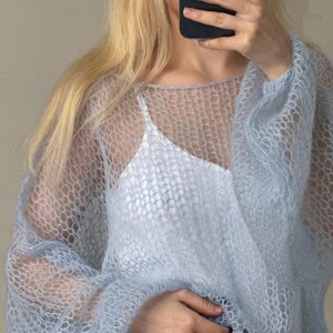 Op de afbeelding: Een lichtblauwe, open gebreide trui met lange mouwen. De trui wordt over een witte camisole gedragen. De textuur van de trui is zichtbaar en de algehele stijl is luchtig en delicaat. De mouwen zijn bij de pols verzameld.
