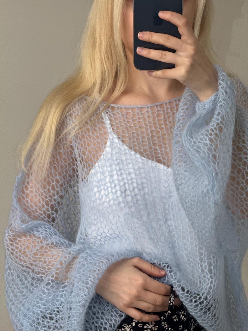 Op de afbeelding: Een lichtblauwe, open gebreide trui met lange mouwen. De trui wordt over een witte camisole gedragen. De textuur van de trui is zichtbaar en de algehele stijl is luchtig en delicaat. De mouwen zijn bij de pols verzameld.
