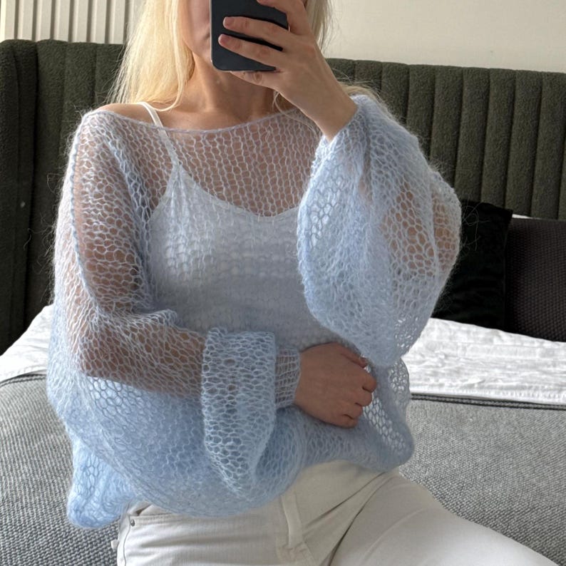 Op de afbeelding: Een lichtblauwe, open gebreide trui met lange, volumineuze mouwen. De trui wordt gedragen over een witte camisole en gecombineerd met een witte broek. Het breiwerk heeft een delicate, luchtige uitstraling.