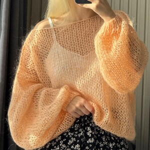 May include: A peach-colored, open-weave sweater with long, voluminous sleeves. The sweater has a relaxed fit and a delicate, airy texture. The garment is paired with a black and white floral skirt.