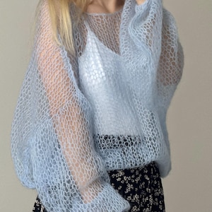 Op de afbeelding: Een lichtblauwe, doorschijnende gebreide trui met lange, volumineuze mouwen. De trui wordt gedragen over een witte camisole en gecombineerd met een zwarte rok met bloemenprint. Het breisel heeft een honingraatpatroon.