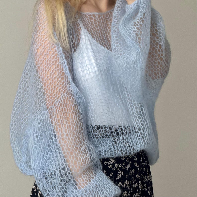Op de afbeelding: Een lichtblauwe, doorschijnende gebreide trui met lange, volumineuze mouwen. De trui wordt gedragen over een witte camisole en gecombineerd met een zwarte rok met bloemenprint. Het breisel heeft een honingraatpatroon.