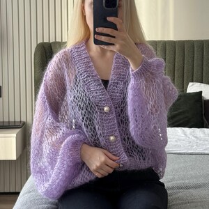 Op de afbeelding: Een lichtpaarse, open geweven cardigan met lange, volumineuze mouwen. De cardigan heeft een V-hals en is vastgemaakt met drie grote, parelachtige knopen. Het kledingstuk lijkt gemaakt van een zacht, pluizig materiaal.