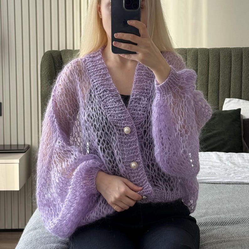 Op de afbeelding: Een lichtpaarse, open geweven cardigan met lange, volumineuze mouwen. De cardigan heeft een V-hals en is vastgemaakt met drie grote, parelachtige knopen. Het kledingstuk lijkt gemaakt van een zacht, pluizig materiaal.