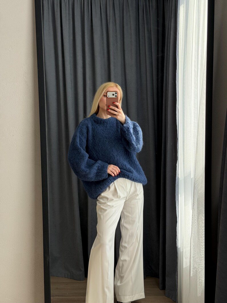 May include: A woman wearing a dark blue, oversized sweater and white wide-leg pants. The sweater has a crew neck and long sleeves. The pants are high-waisted and have a pleated front. The outfit is being reflected in a large mirror.