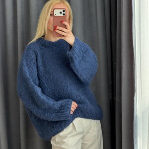 May include: A blue crew neck sweater with long sleeves. The sweater is made of a soft, textured material. The person in the image is wearing white pants. The background is a gray curtain.