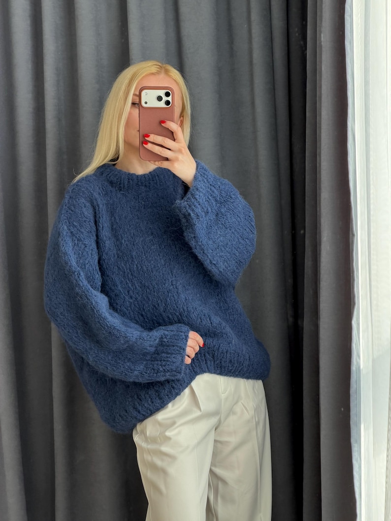 May include: A blue crew neck sweater with long sleeves. The sweater is made of a soft, textured material. The person in the image is wearing white pants. The background is a gray curtain.