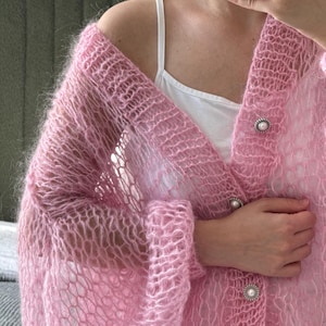 Può includere: Un cardigan rosa chiaro a trama aperta con maniche lunghe. Il cardigan presenta tre bottoni a forma di perla sul davanti. Il capo è realizzato in un materiale morbido e trasparente, creando un aspetto delicato e arioso. Il cardigan è un capo elegante.
