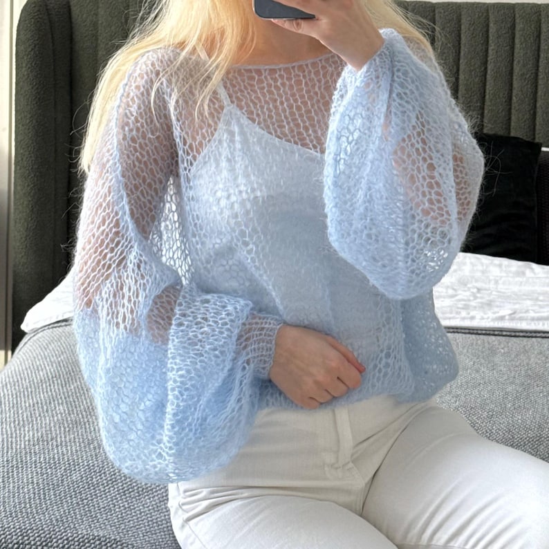 Op de afbeelding: Een lichtblauwe, open gebreide trui met lange, volumineuze mouwen. De trui wordt gedragen over een lichtblauwe camisole en gecombineerd met een witte broek. Het breiwerk heeft een delicate, luchtige uitstraling.