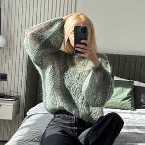 May include: A light green, open-knit sweater with long, voluminous sleeves. The sweater has a slightly sheer appearance and is paired with black pants. The person is holding a phone up to their face.