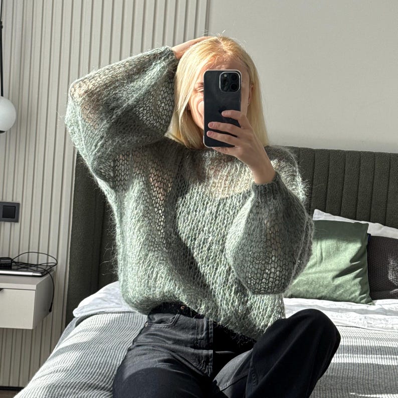 May include: A light green, open-knit sweater with long, voluminous sleeves. The sweater has a slightly sheer appearance and is paired with black pants. The person is holding a phone up to their face.