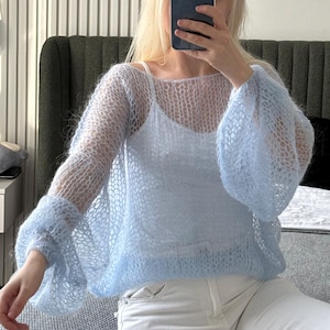 Op de afbeelding: Een lichtblauwe, open gebreide trui met lange, uitlopende mouwen. De trui wordt over een witte camisole gedragen. Het kledingstuk heeft een delicate, luchtige uitstraling. De mouwen zijn volumineus en geven de top een dramatische flair.