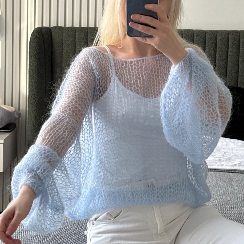 Op de afbeelding: Een lichtblauwe, open gebreide trui met lange, uitlopende mouwen. De trui wordt over een witte camisole gedragen. Het kledingstuk heeft een delicate, luchtige uitstraling. De mouwen zijn volumineus en geven de top een dramatische flair.