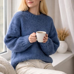 May include: A woman wearing a blue sweater and white pants, holding a white mug. The sweater has a textured appearance. The scene is set near a window with a white vase and decorative dried plants.
