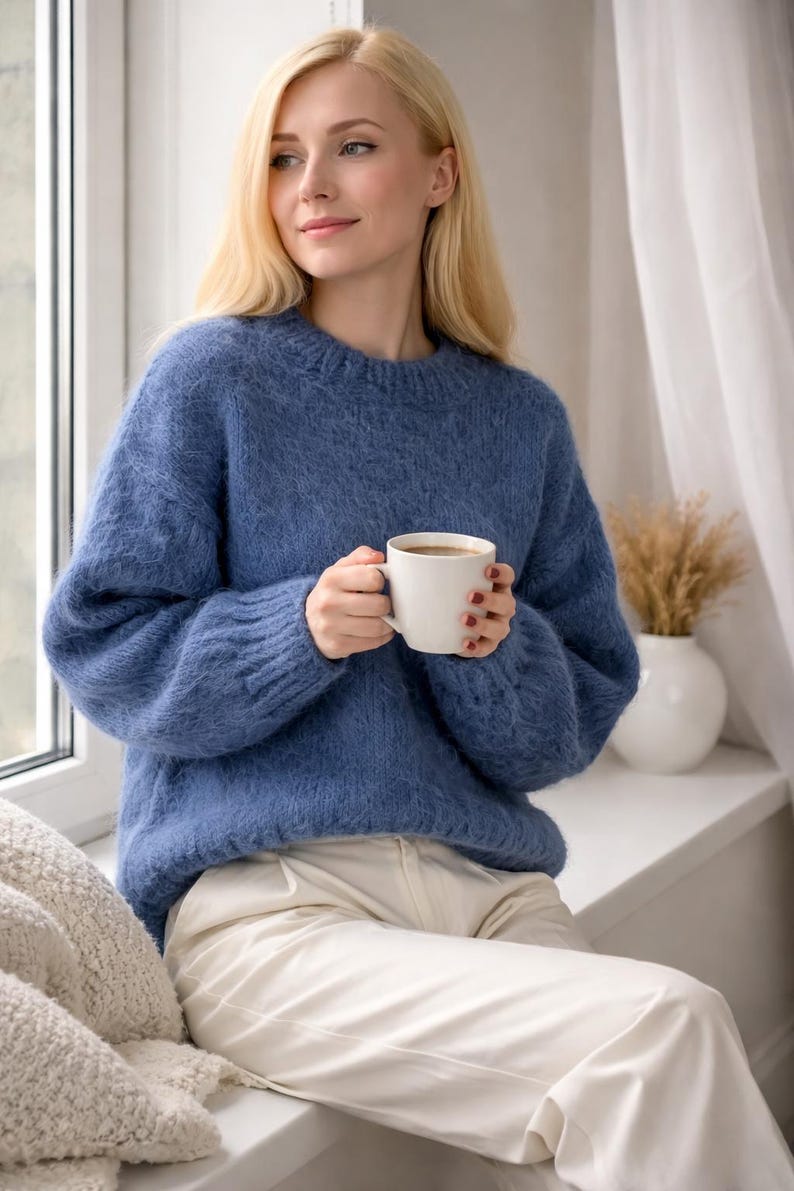 May include: A woman wearing a blue sweater and white pants, holding a white mug. The sweater has a textured appearance. The scene is set near a window with a white vase and decorative dried plants.