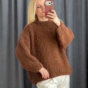 May include: A brown, oversized sweater with a crew neck. The sweater is made of a thick, textured knit material. The person in the image is wearing the sweater with white pants. The sweater appears to be warm and comfortable.