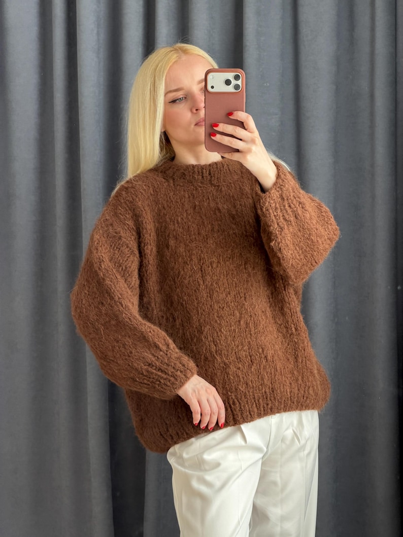 May include: A brown, oversized sweater with a crew neck. The sweater is made of a thick, textured knit material. The person in the image is wearing the sweater with white pants. The sweater appears to be warm and comfortable.