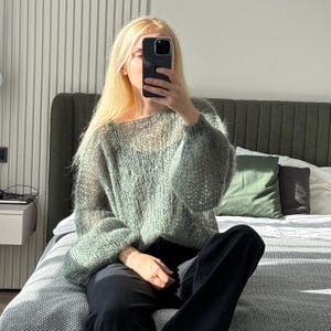 May include: A light green, open-weave sweater with long, puffy sleeves. The sweater is worn with black pants. The person is sitting on a bed with a gray comforter and green pillows. The background includes a headboard and a window.