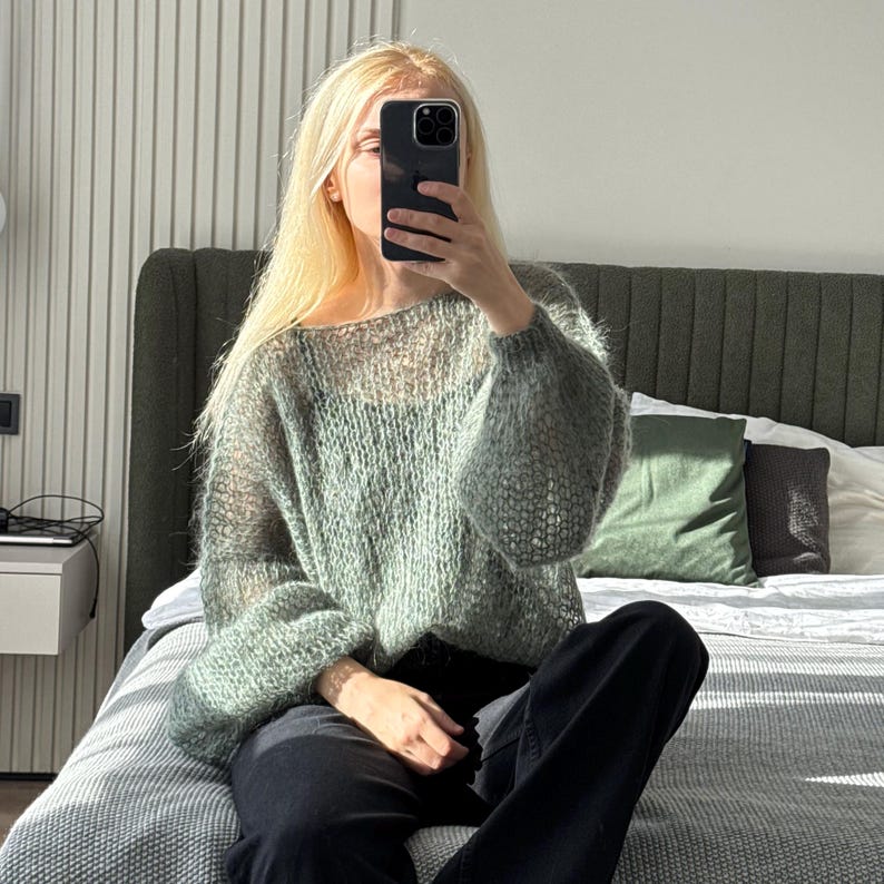 May include: A light green, open-weave sweater with long, puffy sleeves. The sweater is worn with black pants. The person is sitting on a bed with a gray comforter and green pillows. The background includes a headboard and a window.