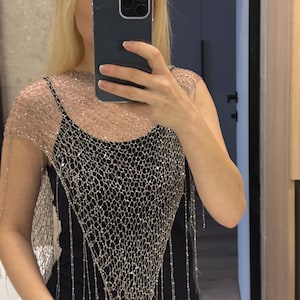May include: A silver mesh top with sparkling embellishments and dangling strands, worn over a black spaghetti strap top. The mesh overlay creates a shimmering effect, adding a touch of glamour to the outfit.