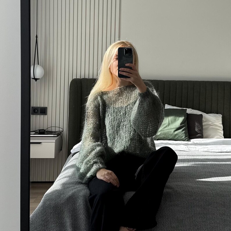 May include: A light green, oversized knit sweater with long, voluminous sleeves. The sweater is paired with black pants. The person is sitting on a bed with a gray comforter and pillows. The room has a modern design with a neutral color palette.