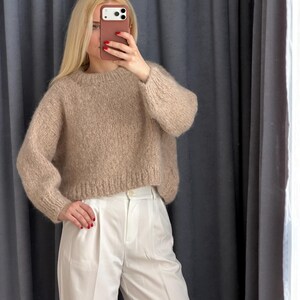 May include: A beige, crew-neck sweater with long sleeves is paired with white, high-waisted pants. The sweater has a textured, knitted appearance. The pants are a crisp white, with a tailored fit. The outfit is simple and elegant.