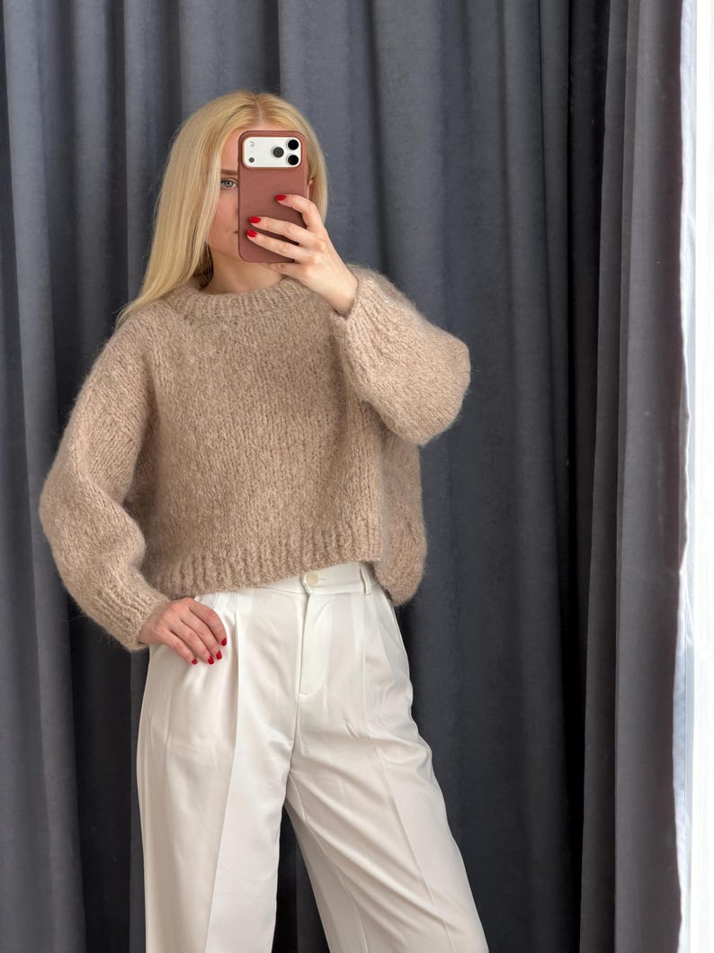 May include: A beige, crew-neck sweater with long sleeves is paired with white, high-waisted pants. The sweater has a textured, knitted appearance. The pants are a crisp white, with a tailored fit. The outfit is simple and elegant.