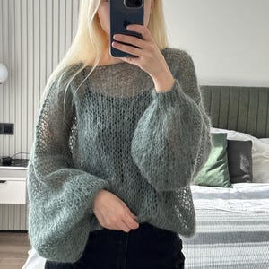 May include: A light green, open-weave sweater with long, voluminous sleeves. The sweater has a round neckline and a loose, airy fit. The sleeves are gathered at the wrist, creating a balloon effect. The material appears soft and slightly fuzzy.