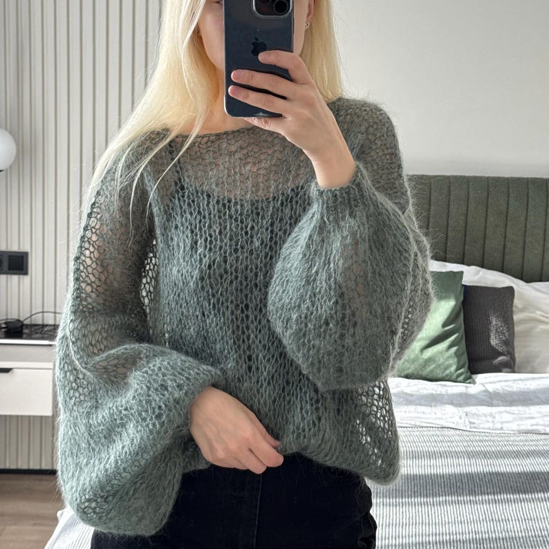 May include: A light green, open-weave sweater with long, voluminous sleeves. The sweater has a round neckline and a loose, airy fit. The sleeves are gathered at the wrist, creating a balloon effect. The material appears soft and slightly fuzzy.