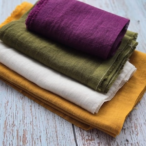 May include: A stack of folded linen fabric in various colors. The colors include mustard yellow, white, olive green, and a deep purple. The fabric has a slightly textured appearance and a fringed edge.