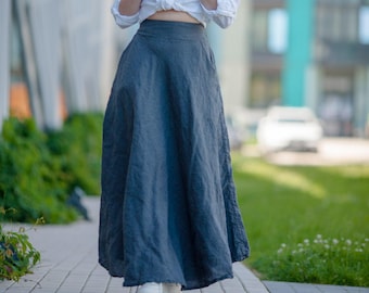 May include: A woman wearing a white button-down shirt tied at the waist, a dark blue maxi skirt, and white sneakers. The skirt has a flowing, A-line silhouette. The background includes a building and greenery.