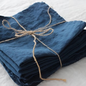 May include: A stack of indigo blue linen napkins tied with natural twine. The napkins have a soft, textured appearance and are neatly folded. The twine is wrapped around the stack and tied in a bow, creating a rustic and elegant presentation.
