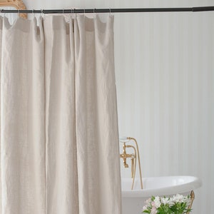 May include: A beige linen shower curtain hangs from a black metal rod in a bathroom. The curtain is made of natural linen fabric and has a simple, elegant design. A white bathtub with gold feet is visible in the background.