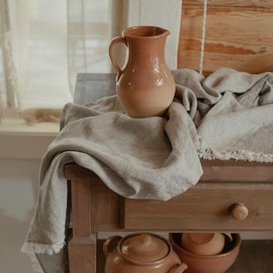 May include: A rustic, earthenware pitcher with a handle sits atop a wooden table draped with a linen cloth. Below, a teapot and bowl are visible. The warm, earthy tones create a cozy, inviting atmosphere. The scene suggests a focus on home decor and kitchenware.
